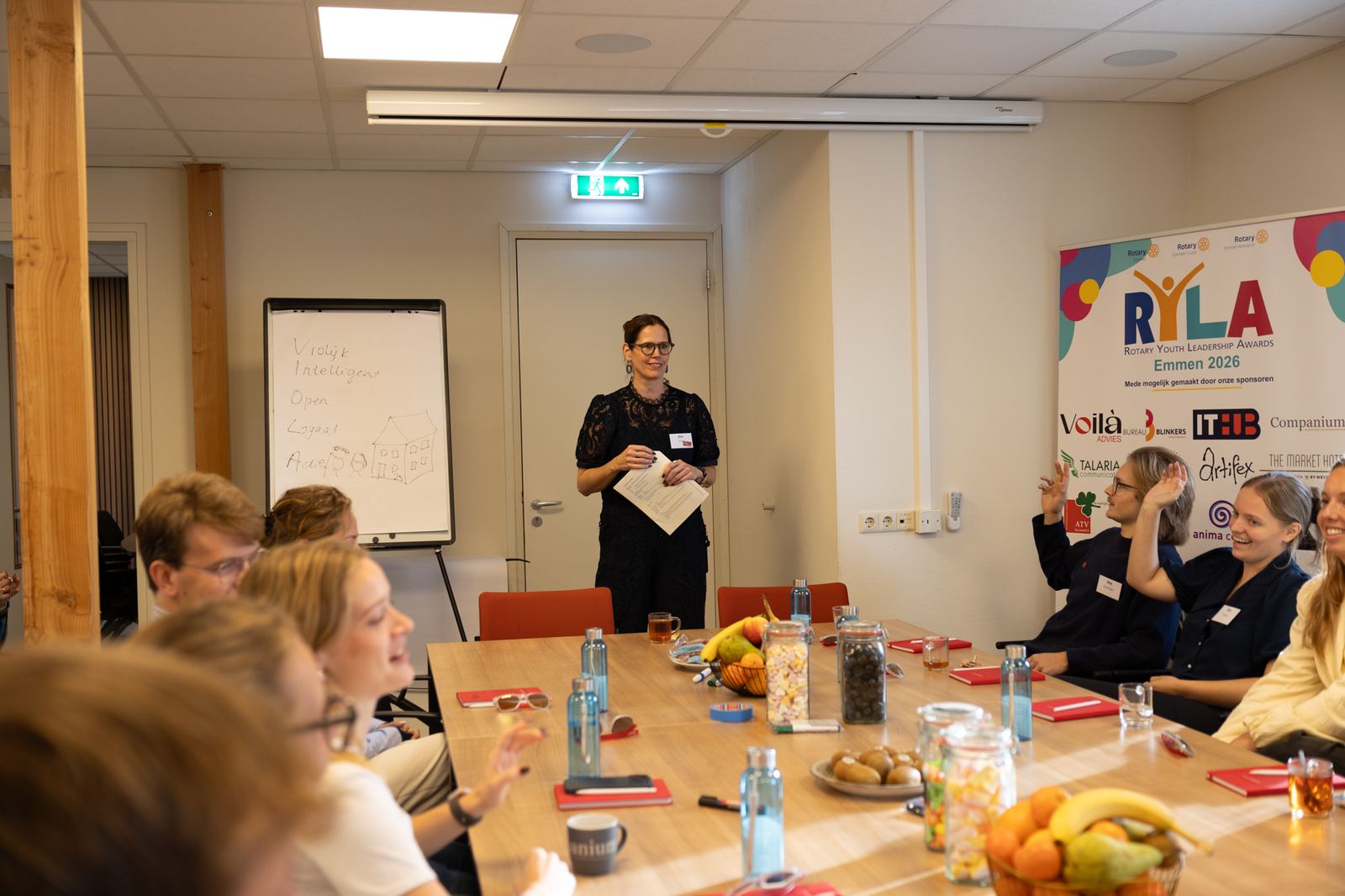 Deelnemers zitten aan een lange tafel en steken hun hand op tijdens een workshop